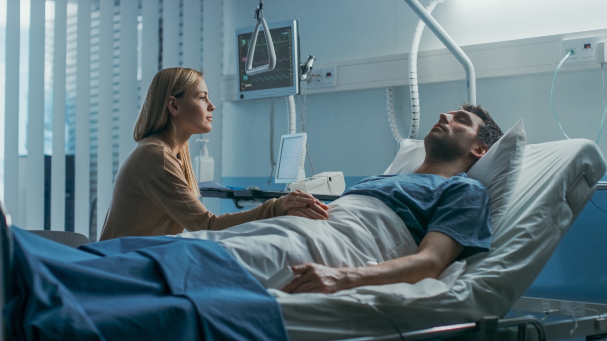 Im Krankenhaus sitzt eine besorgte Ehefrau neben dem Bett, in dem ihr kranker Ehemann liegt. Sie hält seine Hand, redet mit ihm und hofft auf Genesung.