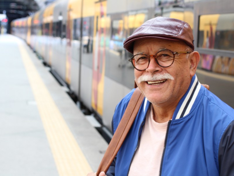 An older man with a blue jacket and a cap is standing in front of a train.