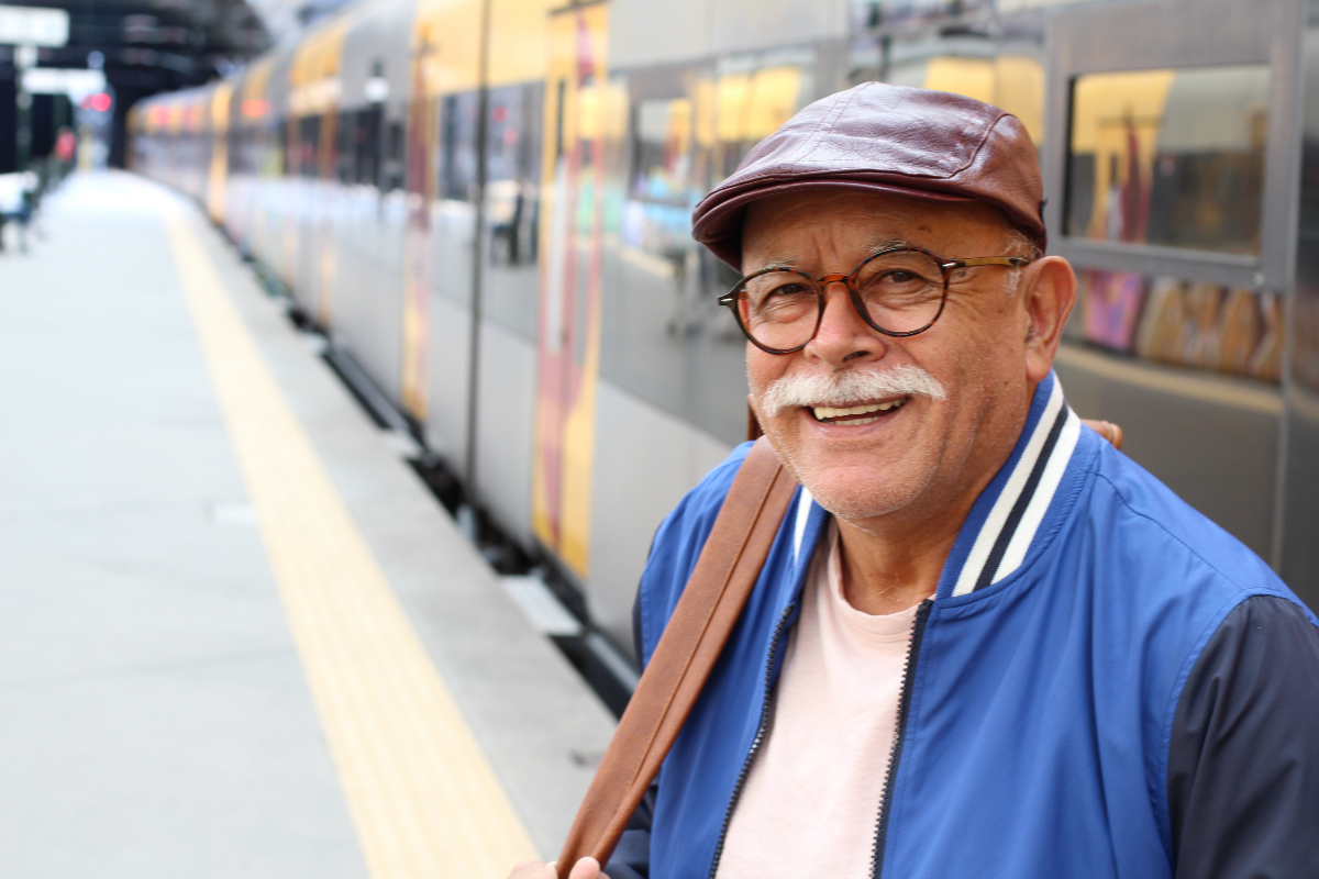 An older man with a blue jacket and a cap is standing in front of a train.