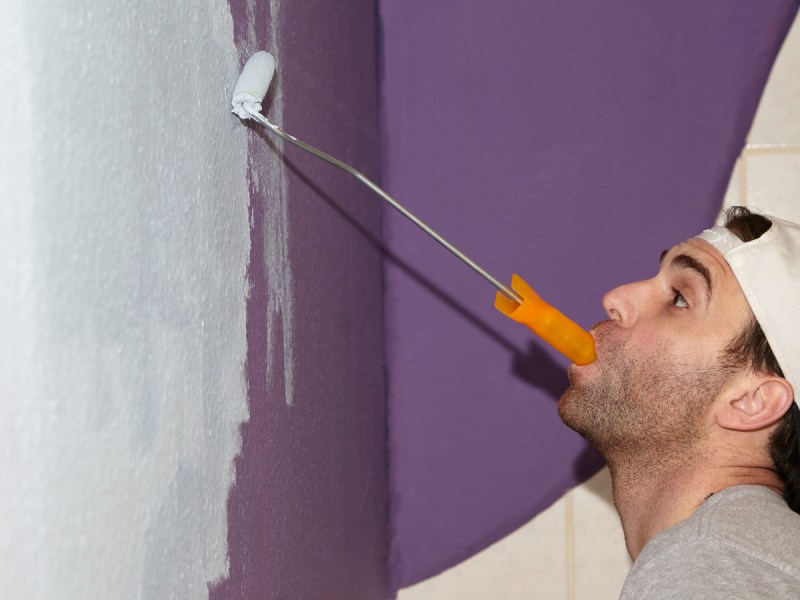 Ein Handwerker streicht eine Zimmerwand und hält dabei den Farbroller mit dem Mund fest.