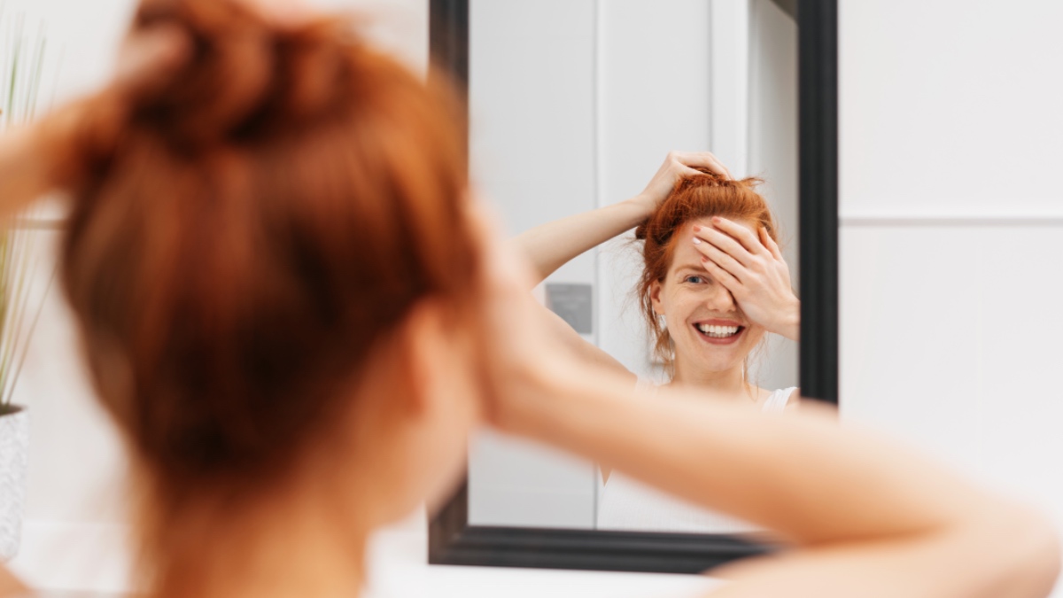 Lachende junge Frau, die sich in einem Spiegel betrachtet, während sie ein Auge mit der Hand abdeckt und sich auf das Spiegelbild konzentriert.