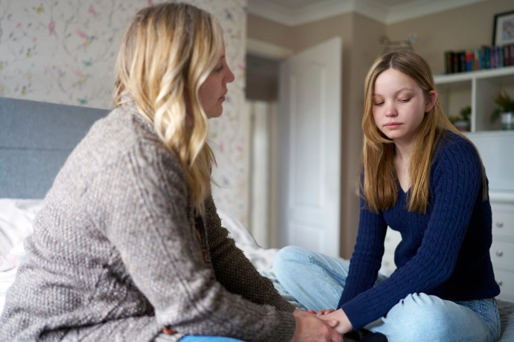 Eine Mutter führt ein ernstes Gespräch mit ihrer Tochter im Teenager-Alter, auf einem Bett sitzend.